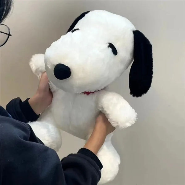 Une personne tient un oreiller en peluche Snoopy blanc avec des oreilles noires sur un fond clair.