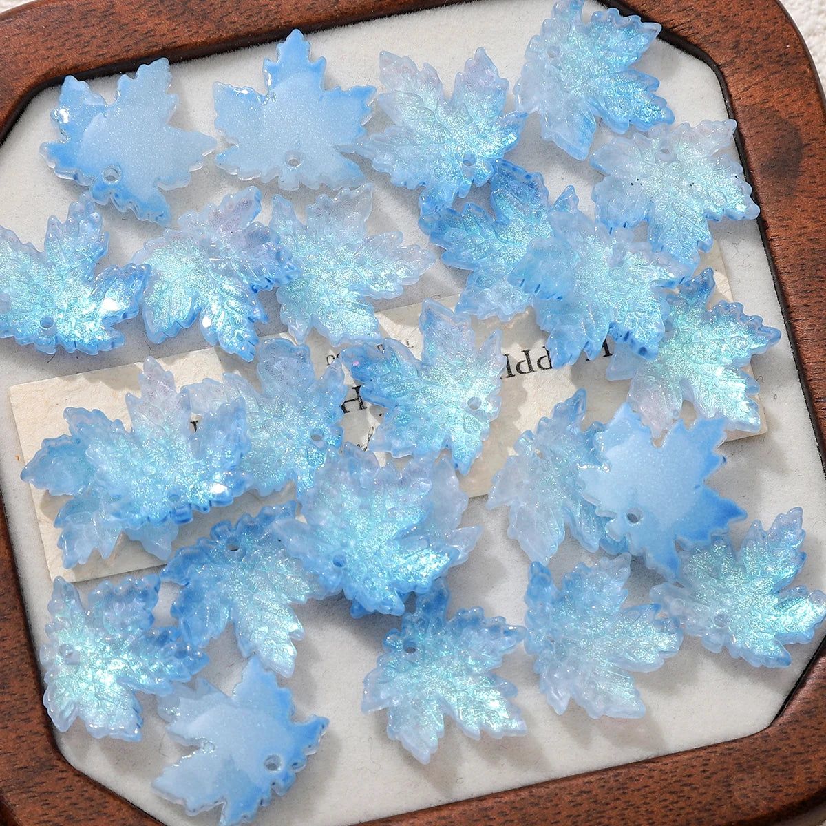 A collection of light blue maple leaf glass beads arranged on a white surface.