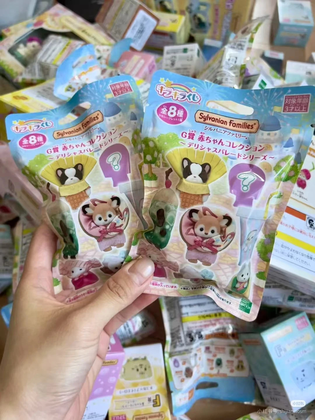 A hand holds a Sylvanian Families blind bag collectible figure package featuring a yellow-eared animal character.