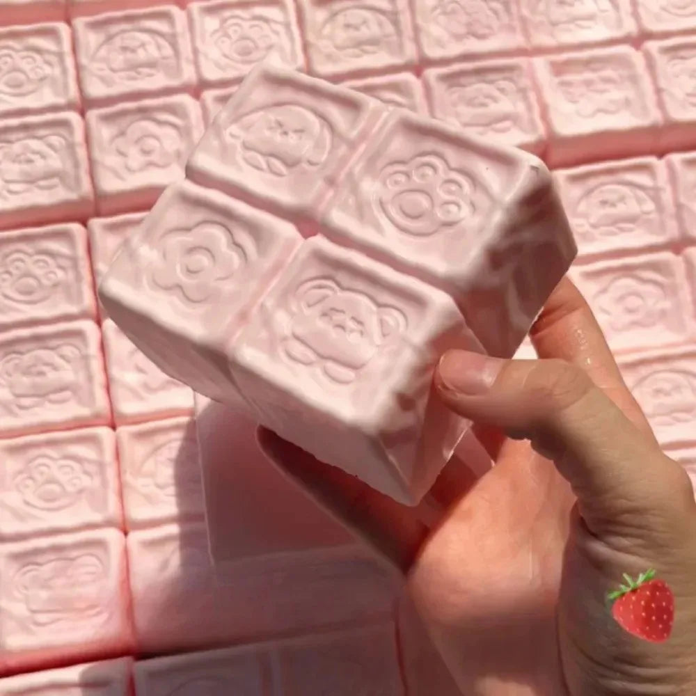 A hand holds a pink strawberry milk squishy stress relief toy with a grid pattern on a pink surface.