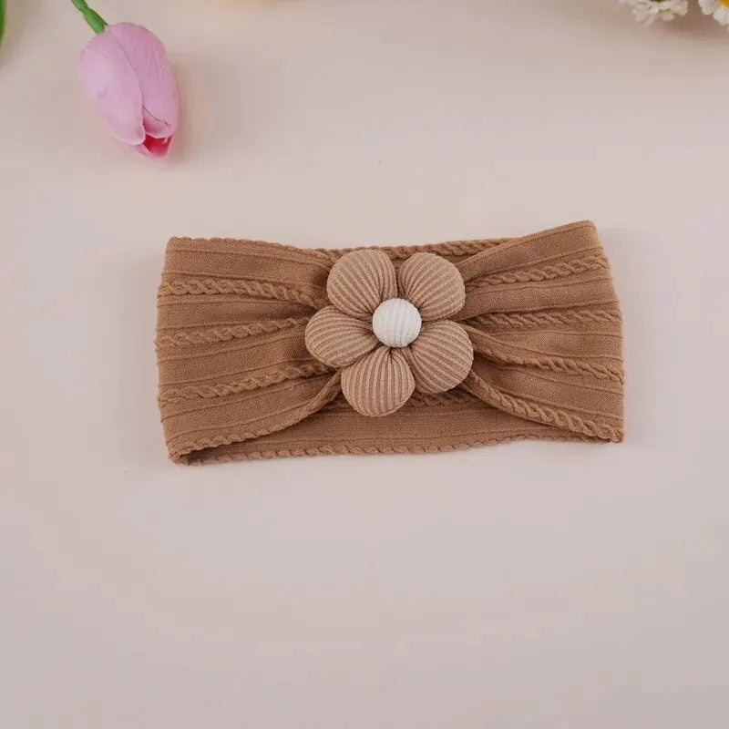 A brown knitted floral elastic baby headband with a white center flower resting on a light pink background.