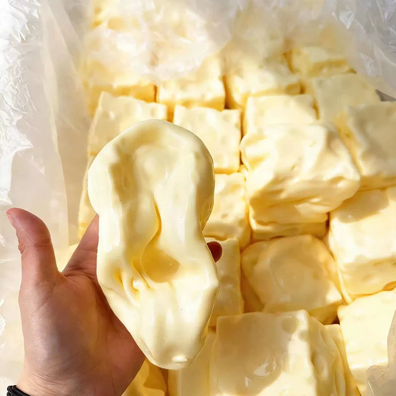A hand holds a soft yellow squishy cheese cube stress ball inside clear plastic packaging.
