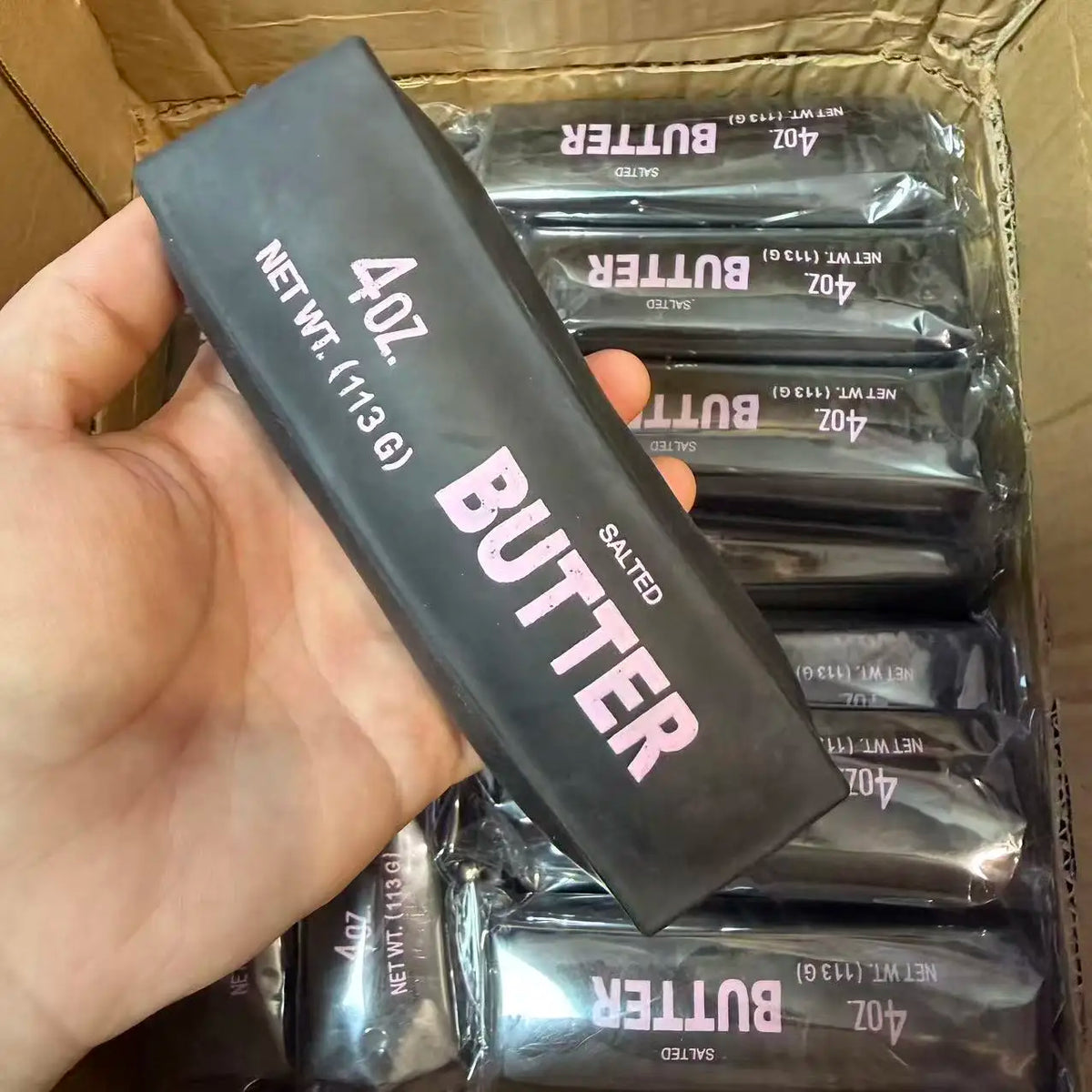A hand holds a black butter stick squishy stress ball with salted butter texture inside its packaging.