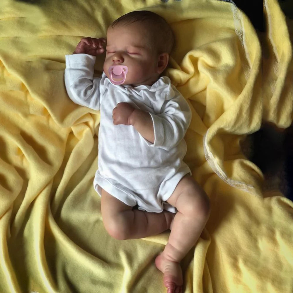 A newborn silicone vinyl reborn baby doll with a cloth body lying on yellow fabric.