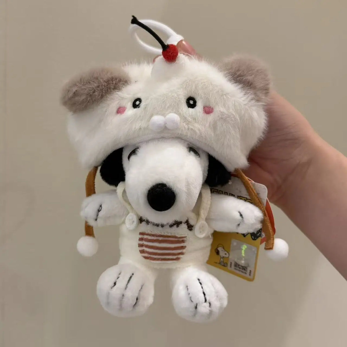 A white Snoopy plush keychain charm with black ears and a red bow is held in a hand.