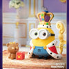 A yellow Minion figure wearing a crown and royal attire sits next to a red box on a patterned rug.