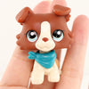 A small, brown and white miniature dog figure wearing a blue scarf is held in a person's hand.
