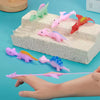 A hand is holding a pink rubber dinosaur toy, with several other colored dinosaurs arranged on a white foam block.