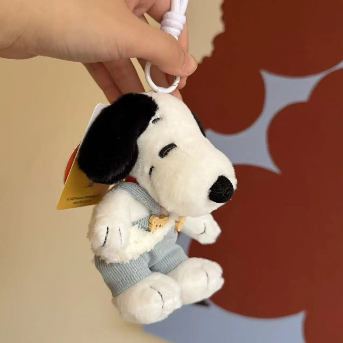 A white Snoopy plush keychain charm is being held by a hand against a blurred orange and blue background.