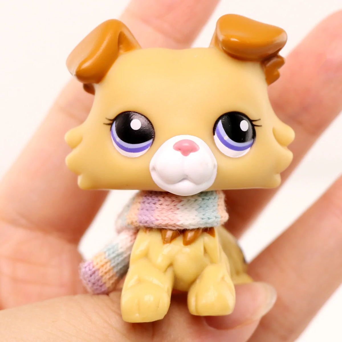 A miniature pet animal figure with light yellow fur, large purple eyes, and a knitted accessory.
