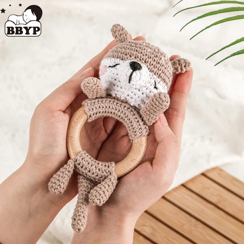 A hand holds a wooden otter rattle and teether ring made of soft beige fabric with a bear face detail.