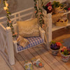 A white miniature dollhouse with furniture and a yellow knitted pillow displayed on a wooden surface.