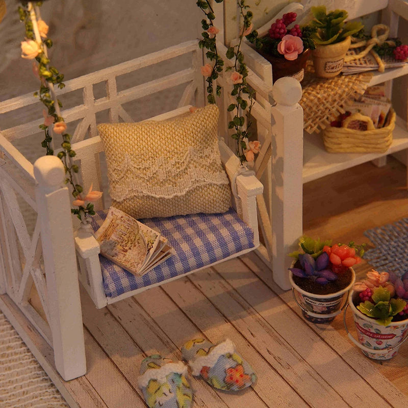 A white miniature dollhouse with furniture and a yellow knitted pillow displayed on a wooden surface.