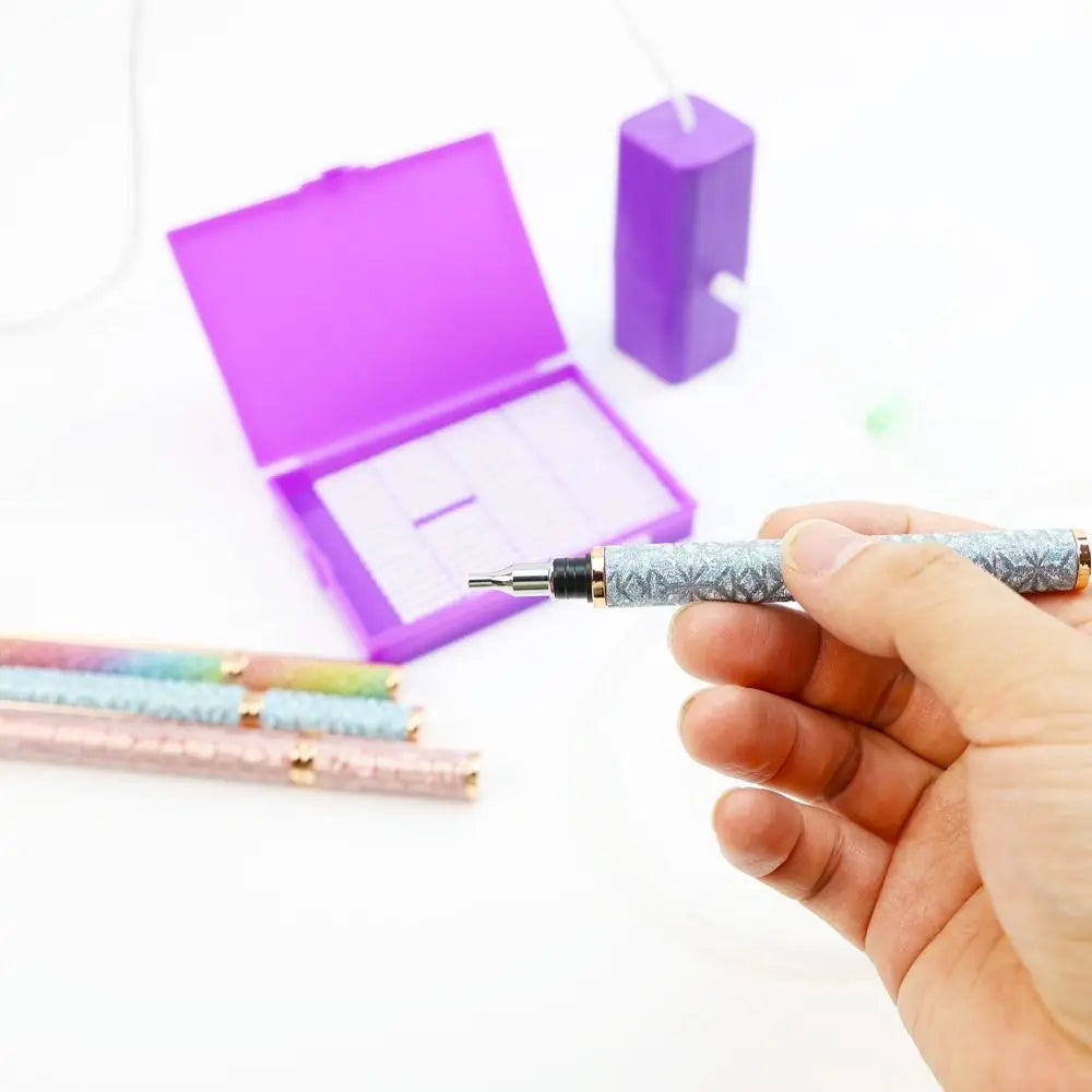 A hand holds an electric diamond painting pen kit with a silver body and purple box in the background.