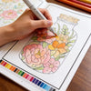 A hand is coloring a detailed botanical illustration of flowers and leaves in a coloring book.