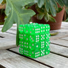A stack of 36 transparent green dice set resting on a wooden surface next to a potted plant.