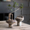 A vintage handcrafted ceramic vase with a spout sits on a weathered wooden surface next to a small green branch.