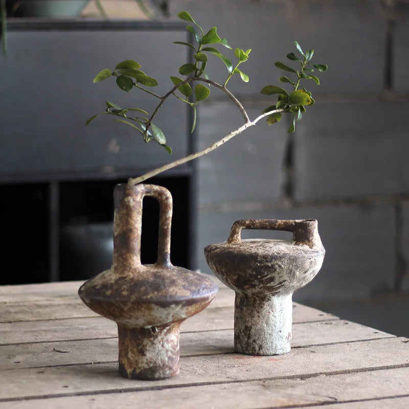 A vintage handcrafted ceramic vase with a spout sits on a weathered wooden surface next to a small green branch.