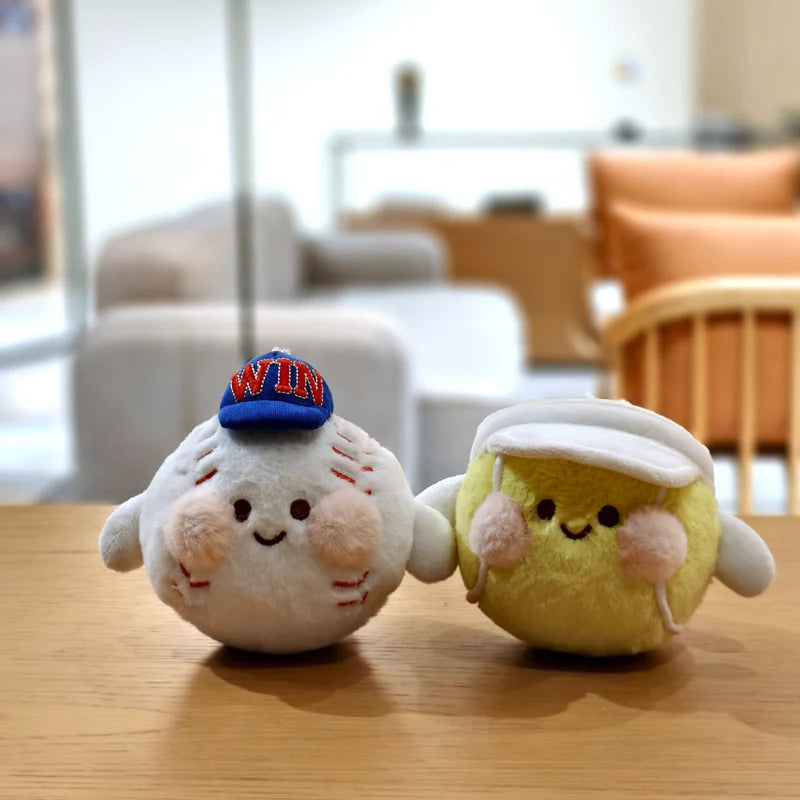 Two fluffy plush charms resembling sports balls, one blue and white with text, and one yellow, resting on a wooden surface.