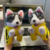 A plush toy resembling a husky dog wearing a yellow outfit is held in front of a laptop.