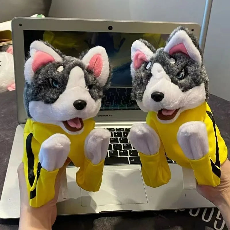A plush toy resembling a husky dog wearing a yellow outfit is held in front of a laptop.