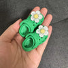 A pair of bright green fashion slippers with white and yellow floral accents are held in a hand.