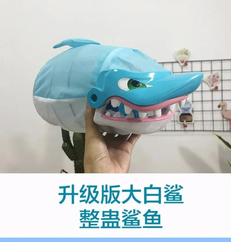 A hand holds a light blue shark-shaped toy with an open mouth showing teeth, used as a prank board game.