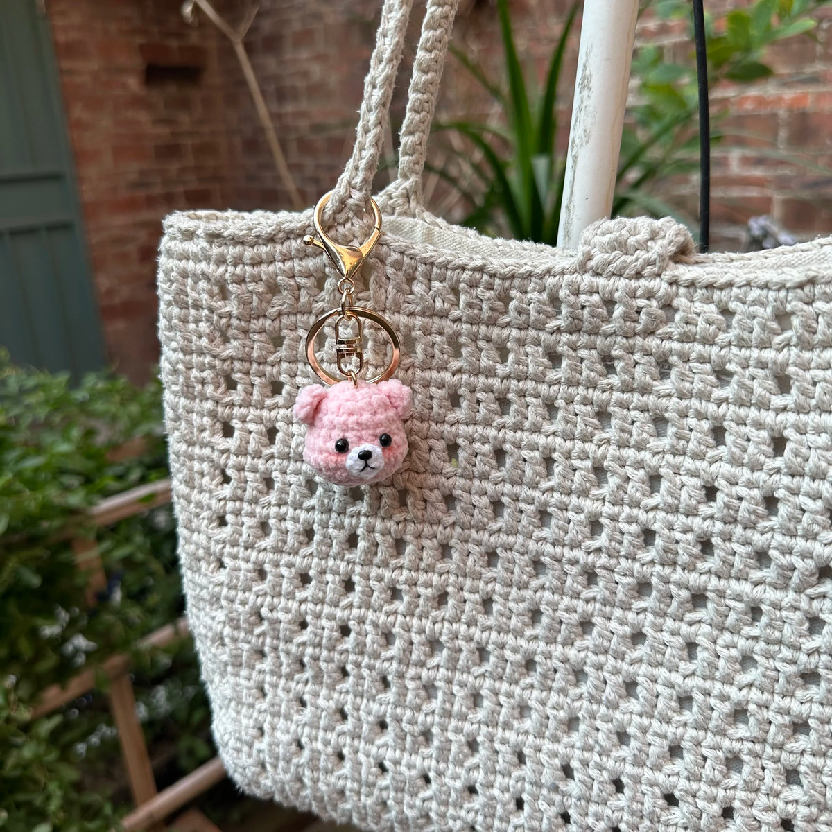 A handcrafted crochet animal keychain in pink, resembling a rabbit or chick, attached to a beige woven bag.