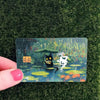A card featuring a cute black cat in the rain amidst green foliage and water ripples.