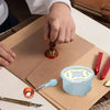 A hand is using a heater and spoon to melt wax for an elegant wax seal kit on brown paper.