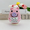 A pink crochet pig ornament with purple details sits on a wooden base, surrounded by yellow and pink tulips.