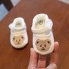 A pair of white soft sole first walker shoes with bear designs are held in a hand on a wooden table.