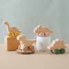A set of wooden dinosaur teether toys in natural wood tones and light pink, resting on a beige surface.