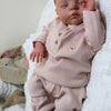 A newborn baby doll with a soft cloth body wearing pink clothing is lying on a white fluffy surface.