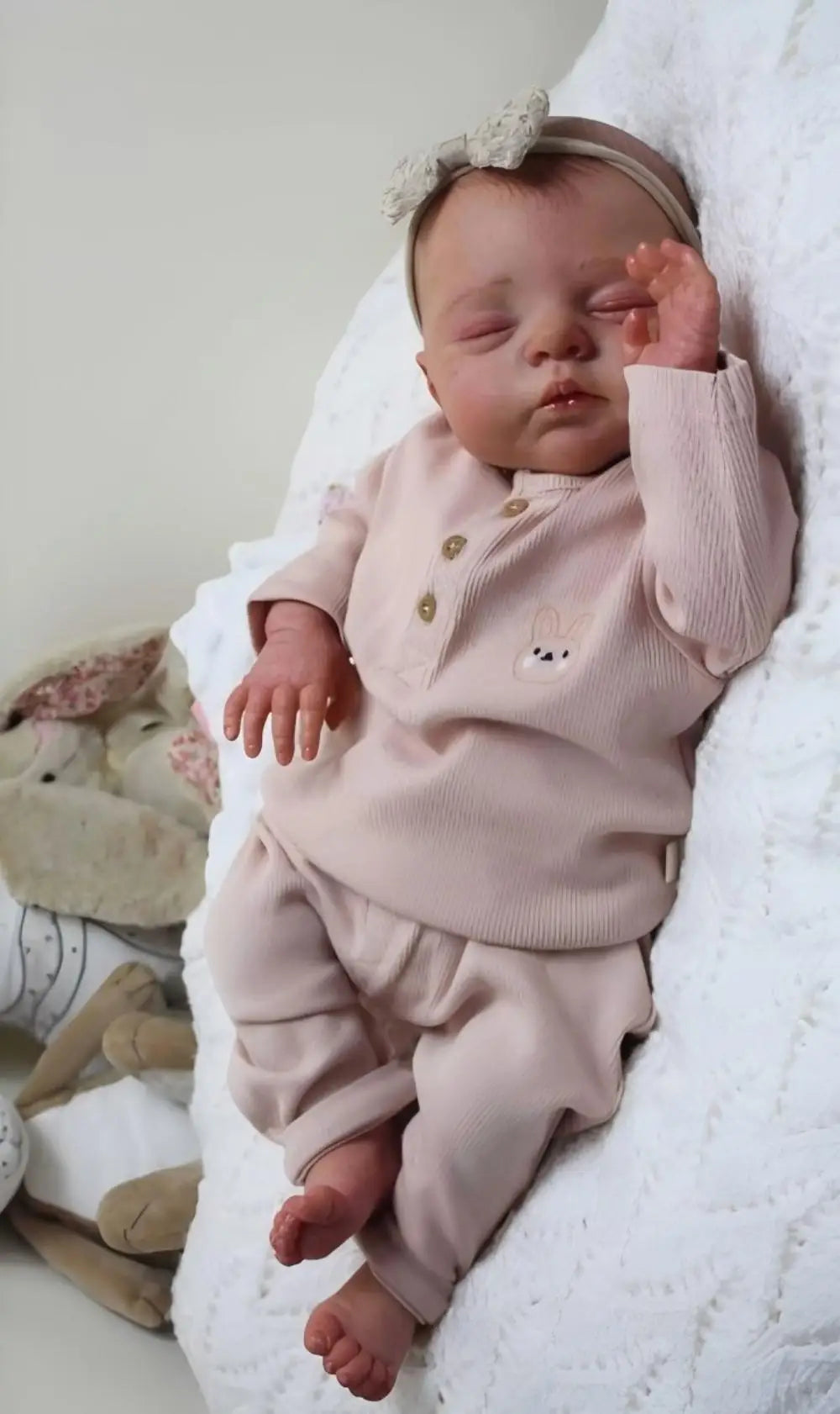 A newborn baby doll with a soft cloth body wearing pink clothing is lying on a white fluffy surface.