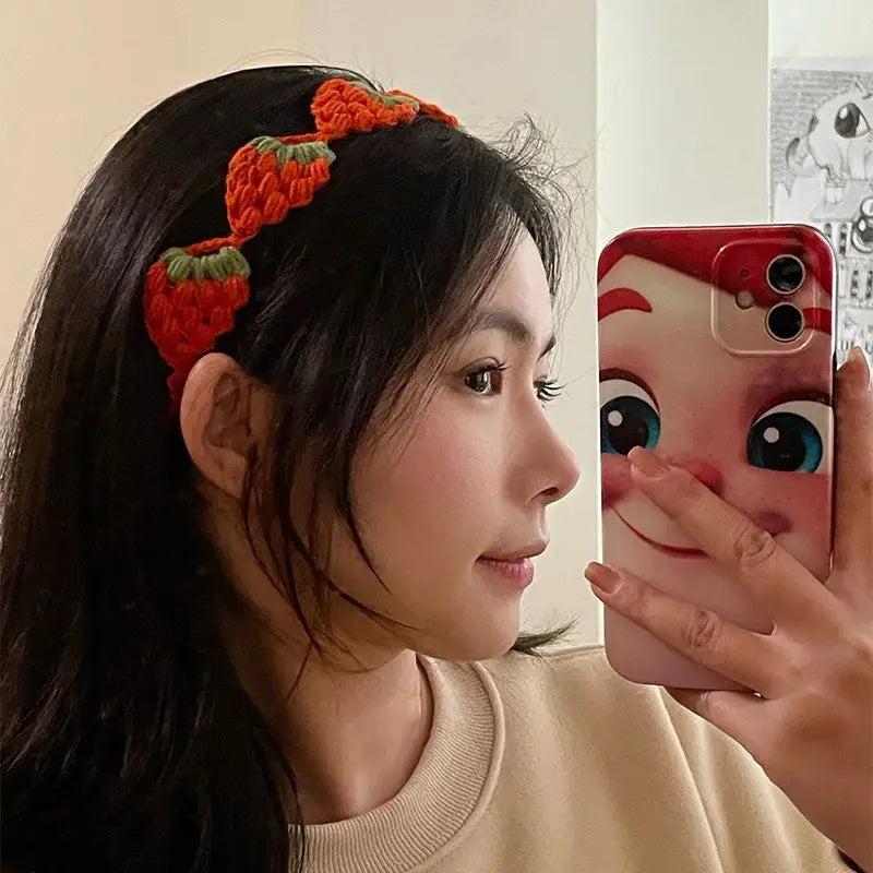 A woman wearing a red strawberry crochet headband while taking a selfie with a smartphone.