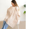 A woman is wearing a beige breathable muslin nursing cover with an adjustable neckline.