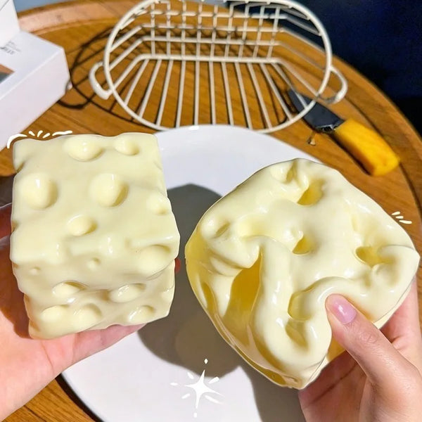 A hand holds a large, yellow squishy cheese cube fidget toy on a white plate next to a wire mesh tool.