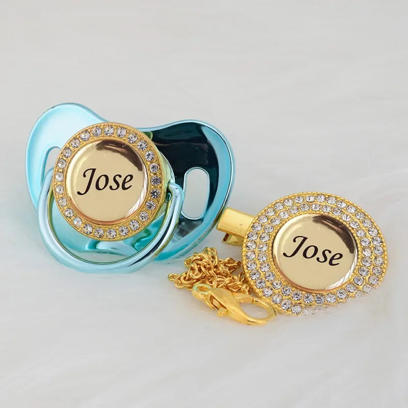 A personalized crystal pacifier and clip gift set featuring a light blue silicone pacifier and a gold-toned clip with a clear
