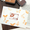 A vintage craft oil-based ink pad in ochre yellow and cocoa brown next to a piece of paper featuring butterfly designs.