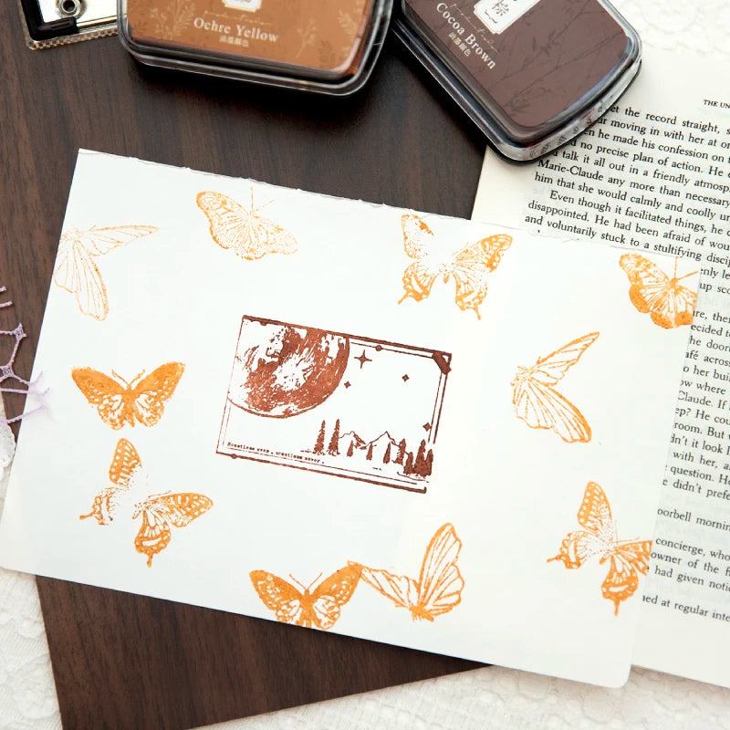 A vintage craft oil-based ink pad in ochre yellow and cocoa brown next to a piece of paper featuring butterfly designs.