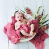 A baby is wrapped in a pink knit wrap with a pearl headband, lying on a light wooden surface surrounded by greenery.