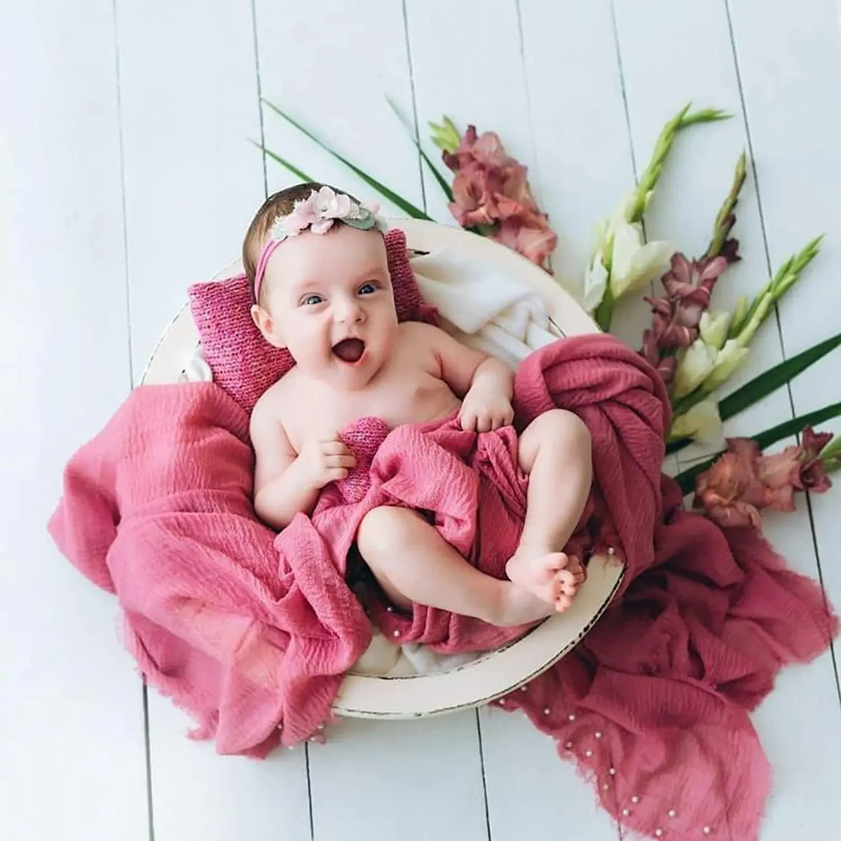 A baby is wrapped in a pink knit wrap with a pearl headband, lying on a light wooden surface surrounded by greenery.