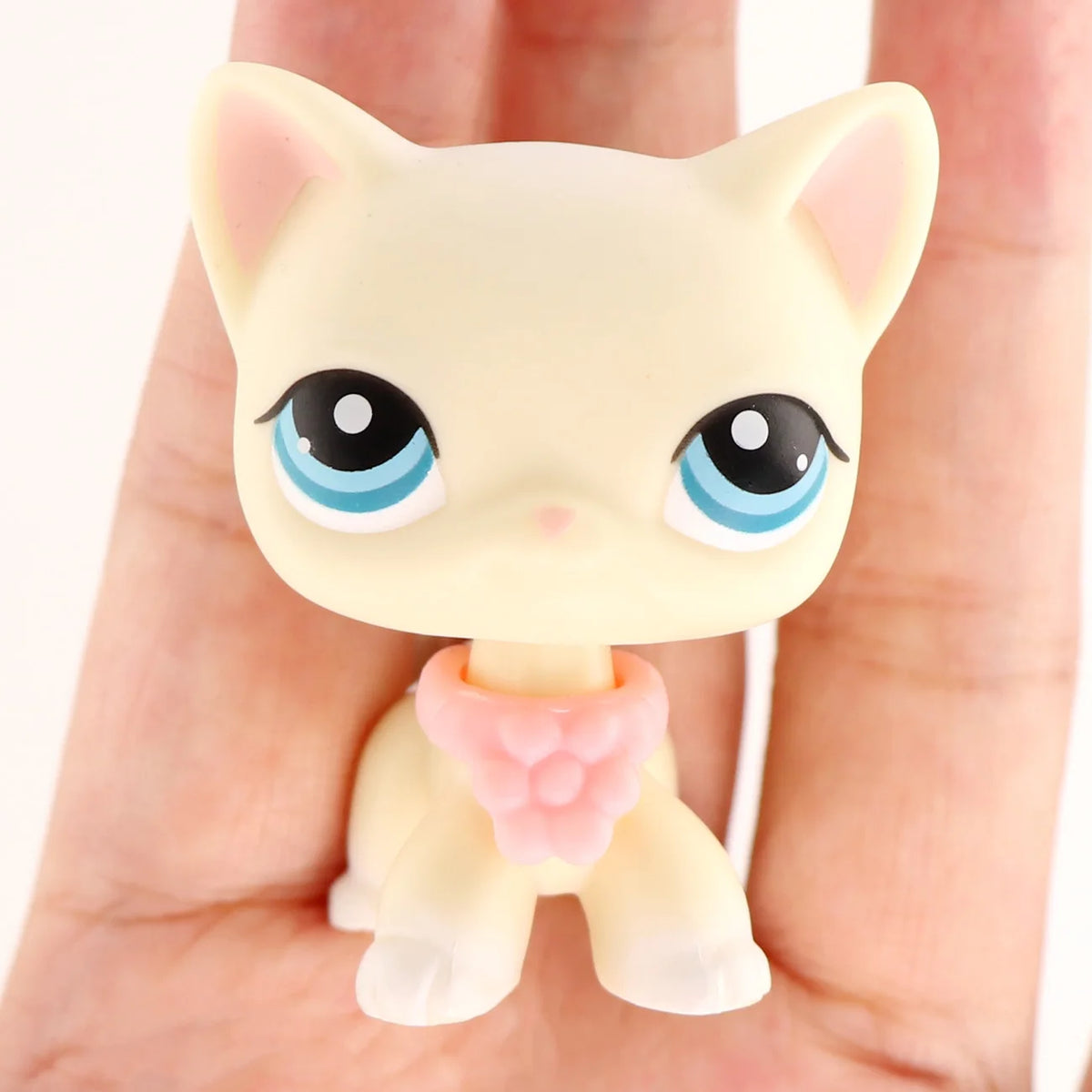 A small, light yellow miniature cat figure with large blue eyes and a pink accessory, held in a person's hand.