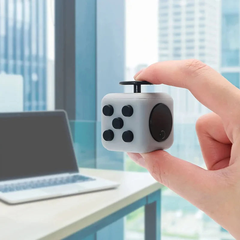 A hand holds a gray fidget cube with magnetic buttons and a rotating joystick against a blurred office window background.