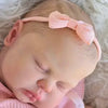 A close-up of a lifelike reborn baby doll with hand-painted skin wearing a pink outfit and headband.