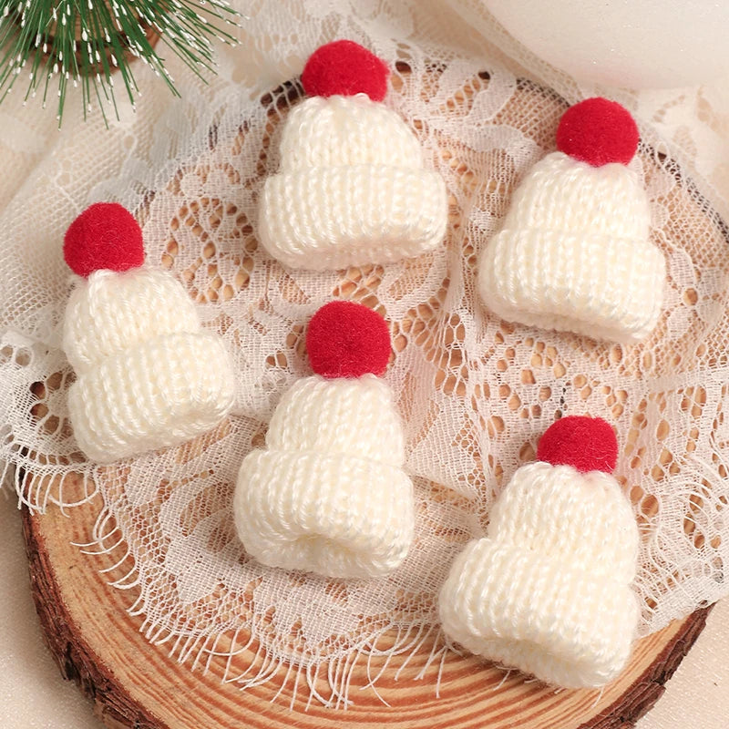 A set of mini knitted white hats with red pom-poms displayed on a lace fabric next to a pine branch.