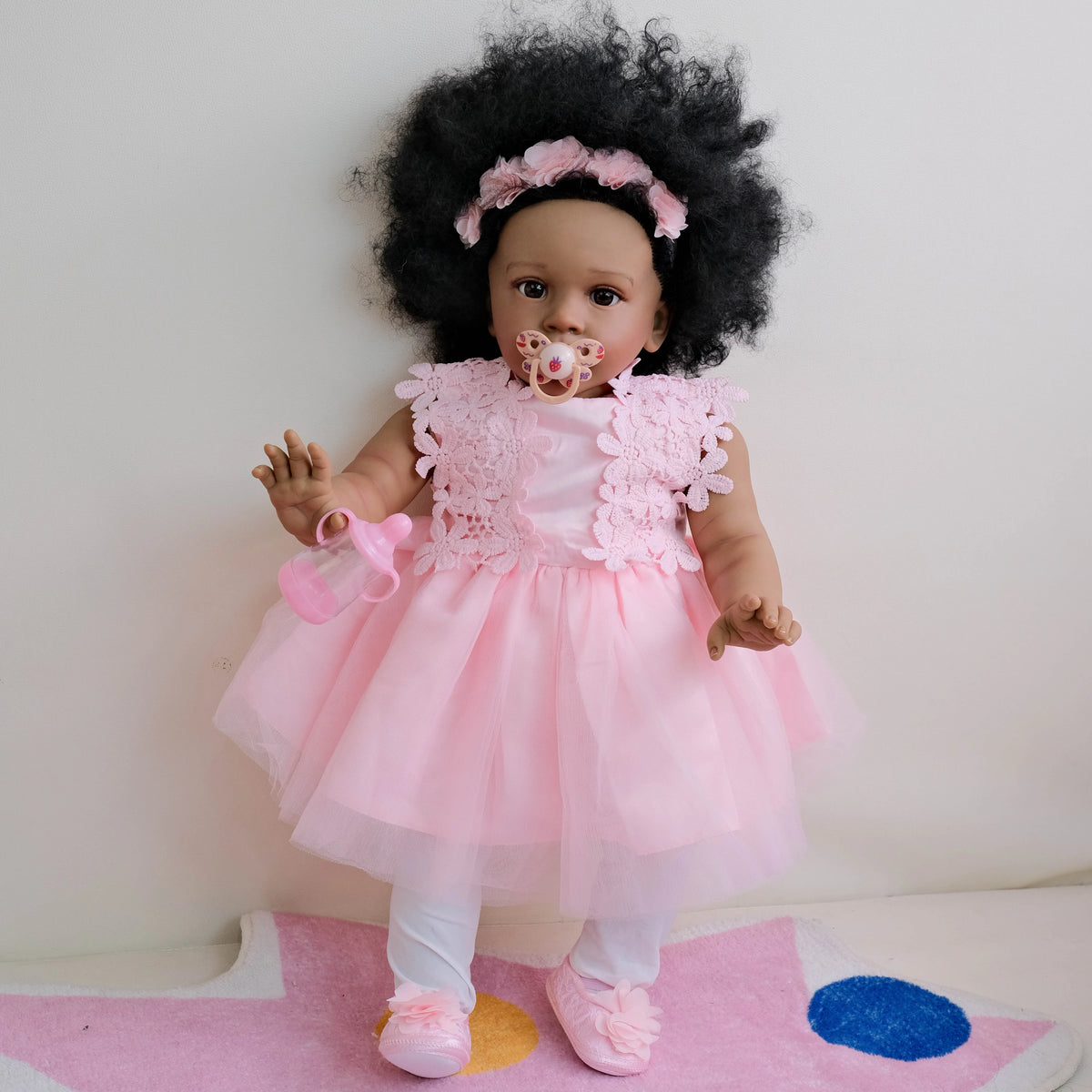 A lifelike reborn toddler doll with hand-rooted black hair and a pink dress sitting on a pink rug.