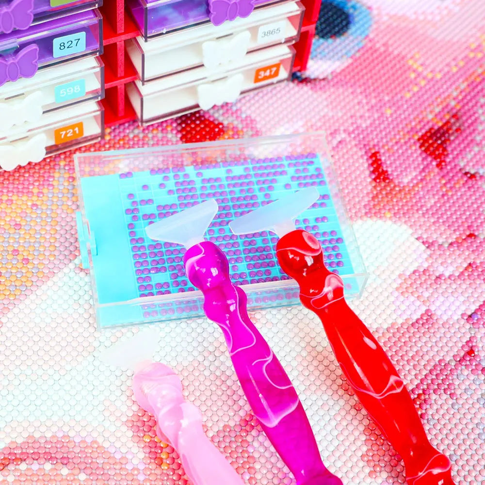 A collection of diamond art pens in various colors and tips displayed on a pink textured surface.