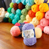 A pile of pre-dyed milk cotton yarn in various shades of pink, yellow, green, and blue is displayed on a wooden surface.